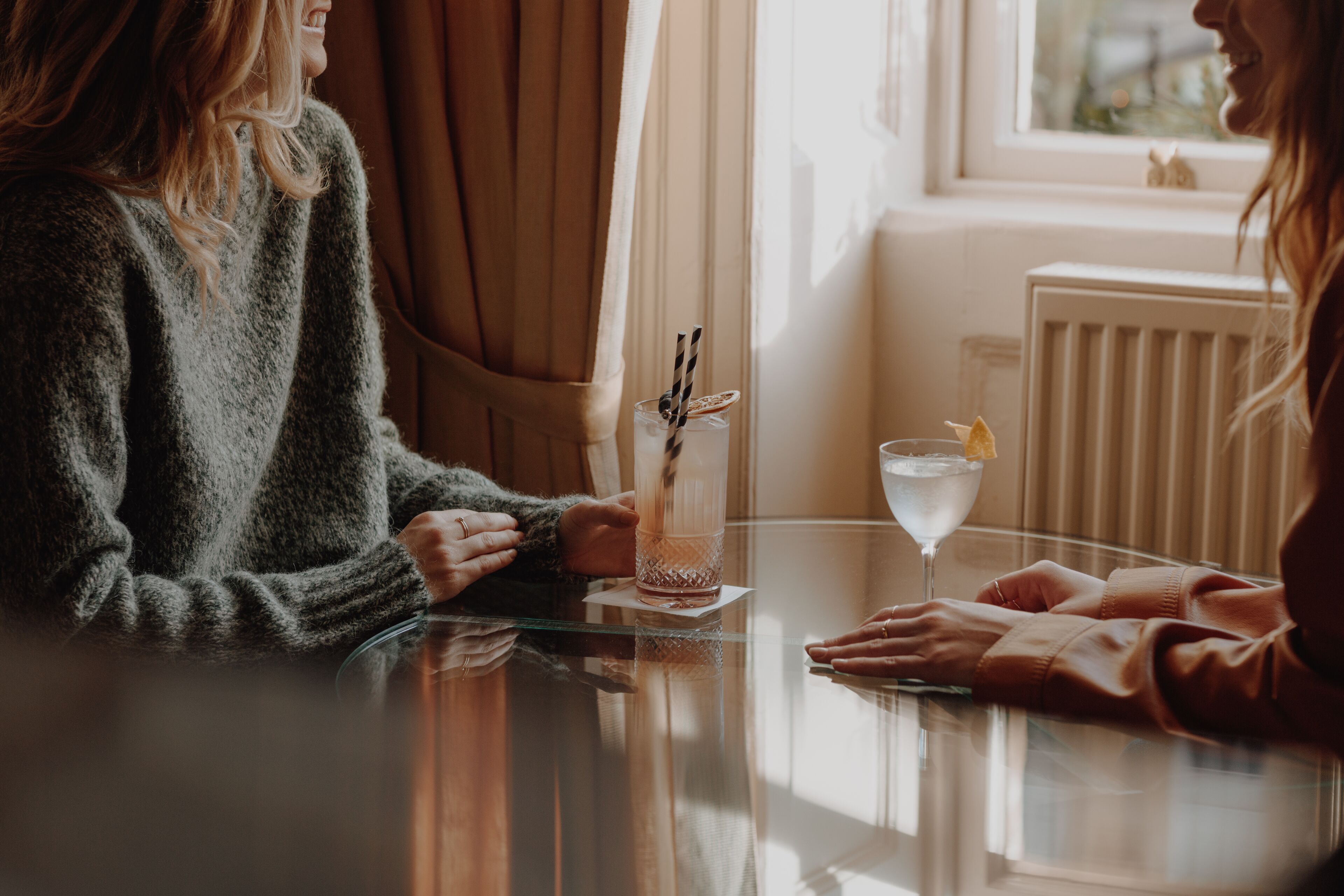 Zwei Personen sitzen an einem Tisch mit Getränken, ein Fenster mit Vorhängen und ein Heizkörper im Hintergrund.