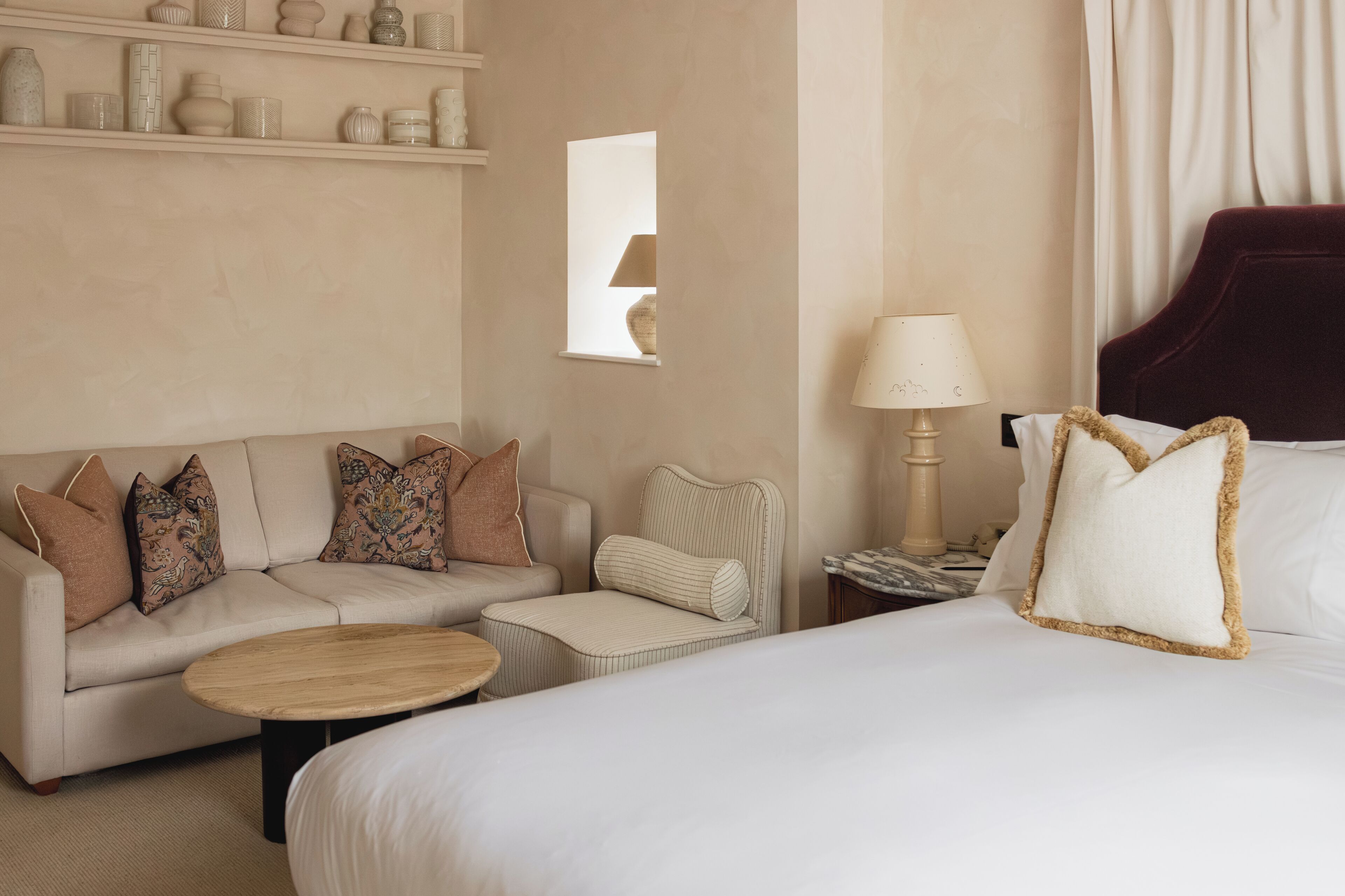 A hotel room with a bed, a sofa, a small table, a lamp, and a shelf with decorative items.