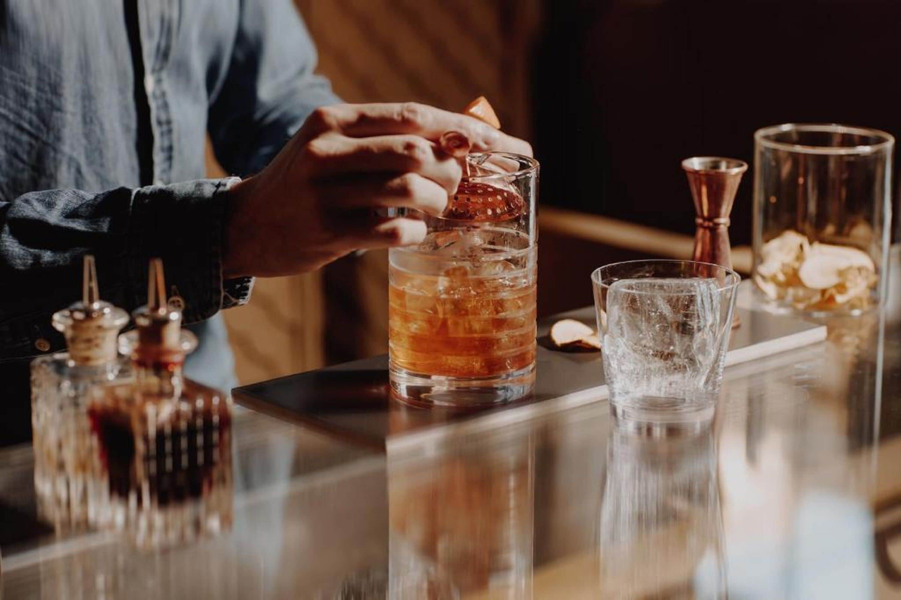 Un ambiente de bar con una persona sirviendo una bebida en un vaso, cubitos de hielo y diversas herramientas de bar.