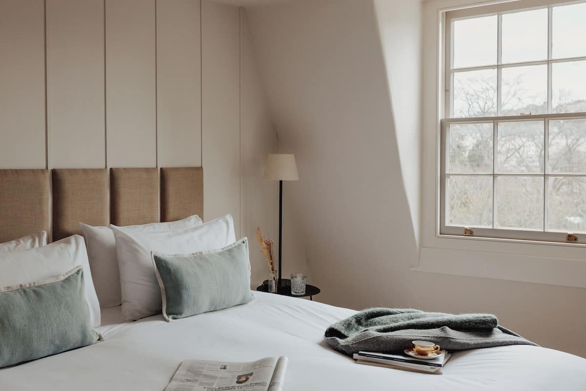 A neatly arranged bedroom with a bed, pillows, a bedside table with a lamp, and a window with a view.