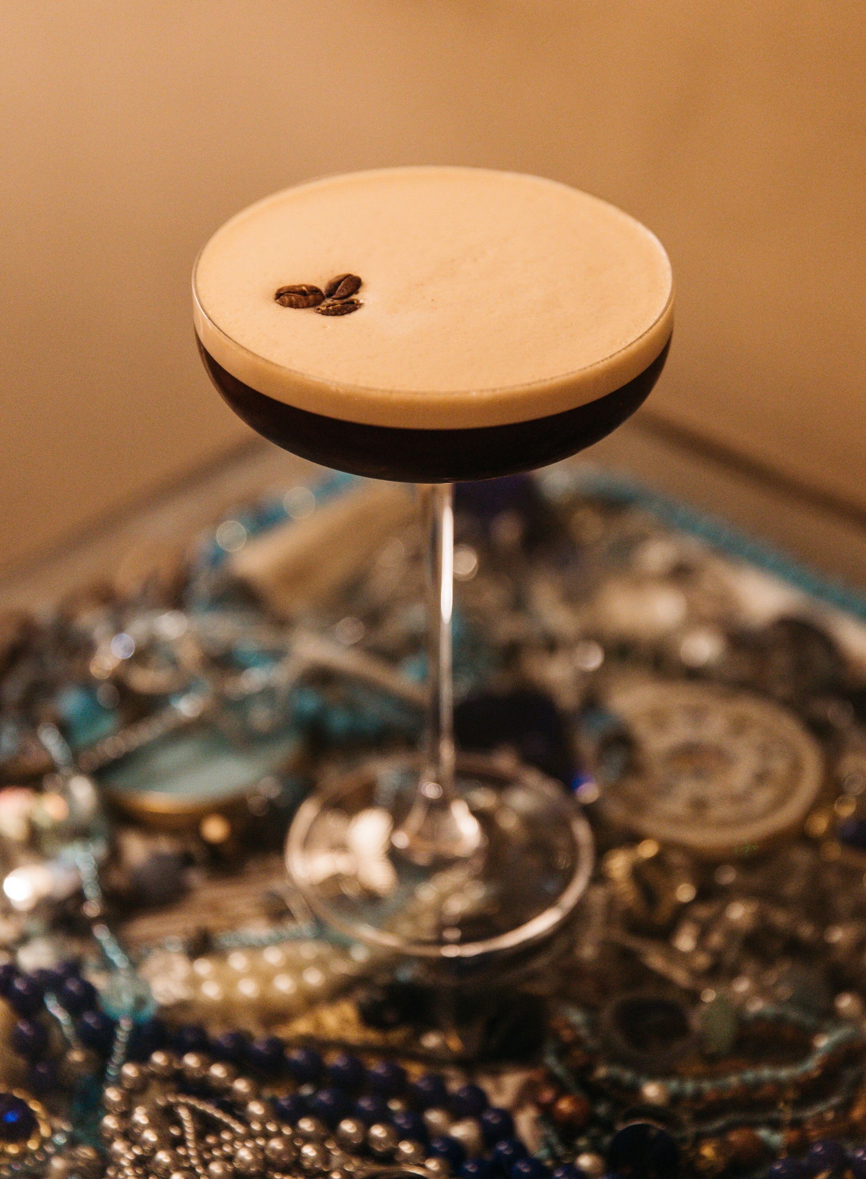 A cocktail glass with a dark liquid and a single coffee bean on the rim, placed on a table with a glass top.