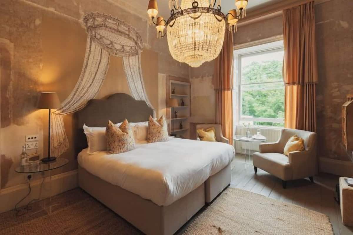A bedroom with a large bed, a chandelier, a chair, a small table, and a view of greenery outside.