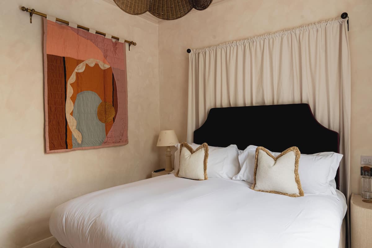 A neatly made bed with white linens and decorative pillows, a bedside lamp, and a wall hanging.