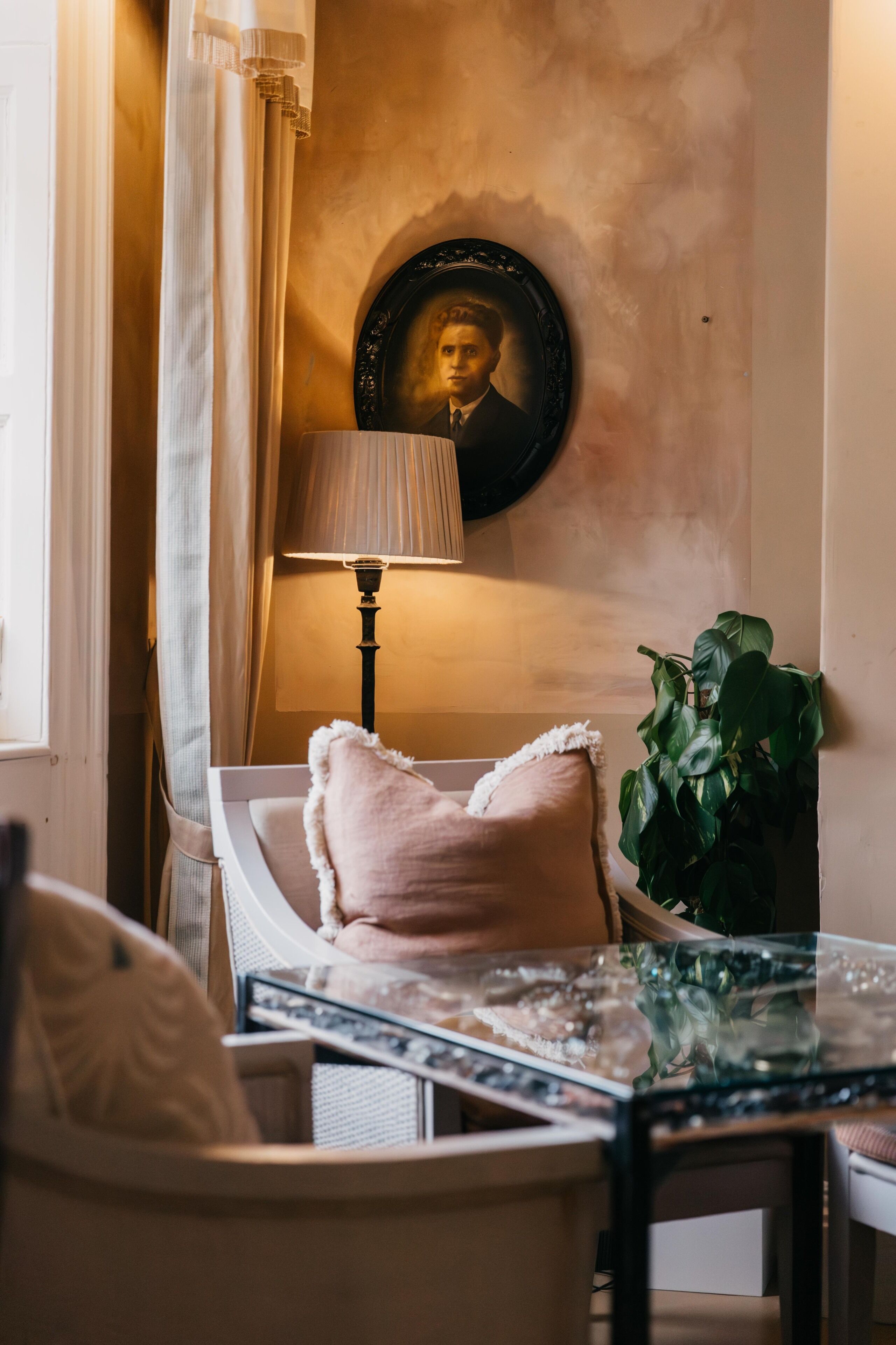A cozy room with a white armchair, a glass table, a lamp, a framed portrait, and a potted plant.