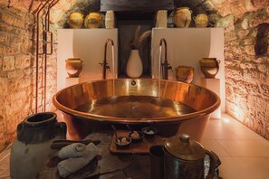 Una habitación con paredes de piedra, una bañera de cobre grande, un florero blanco y varias macetas de cerámica en estantes.