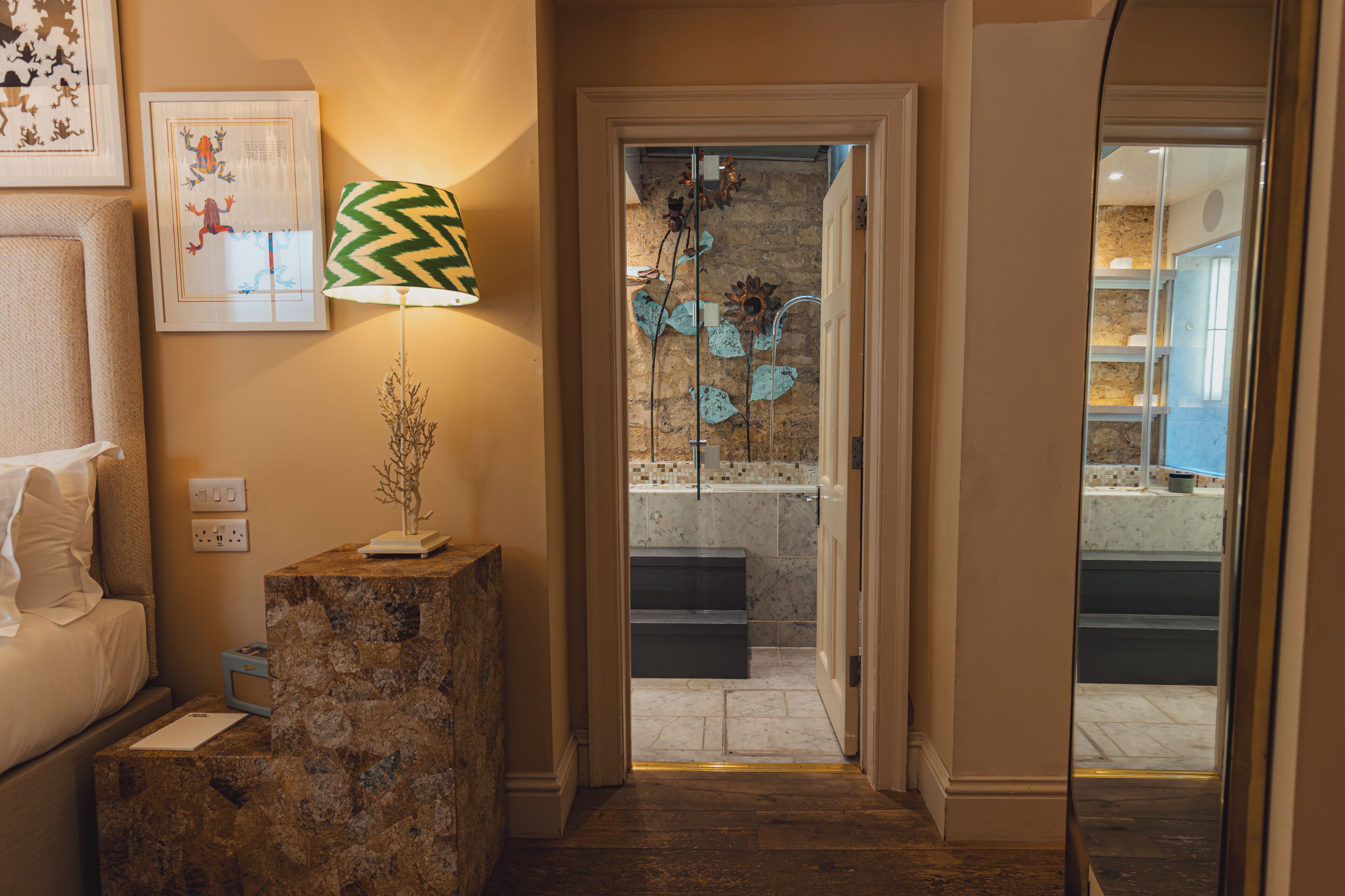 A hotel hallway with a stone pedestal, a lamp, and a door leading to a bathroom with a glass shower and decorative flowers.