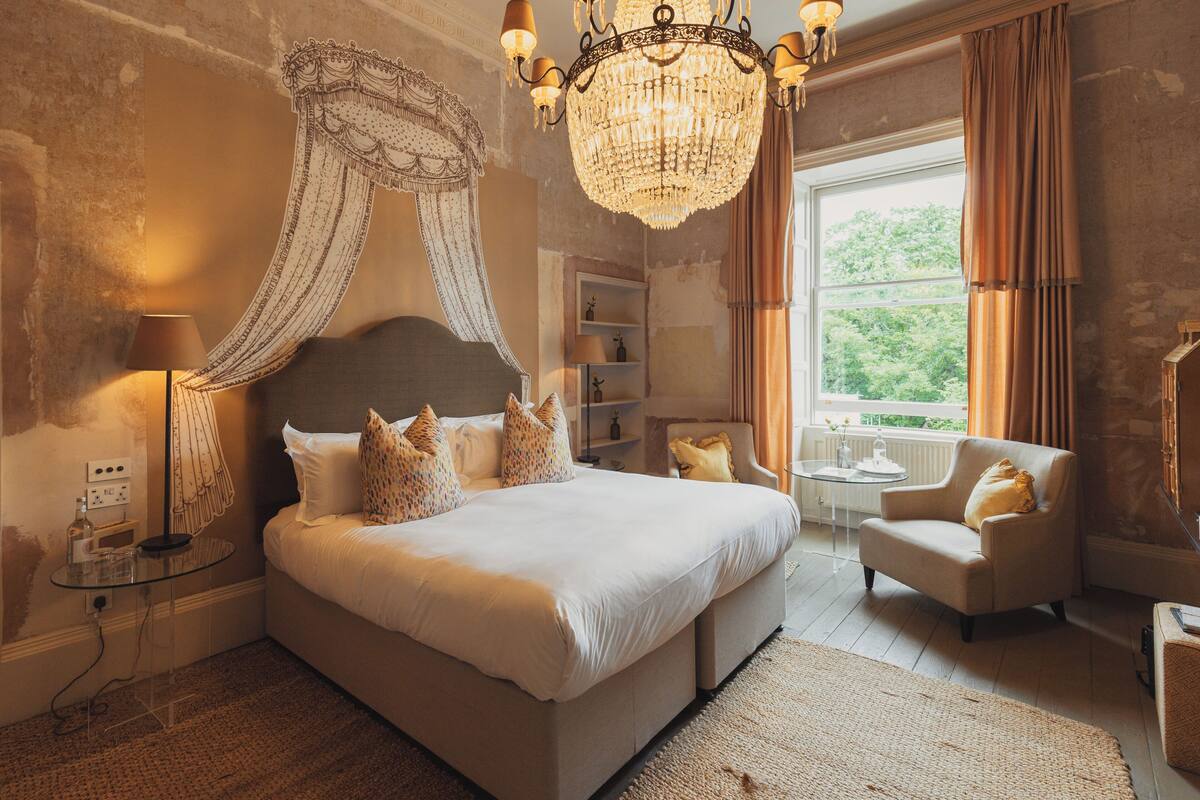 A spacious bedroom with a large bed, a chandelier, a chair, and a view of the outdoors.