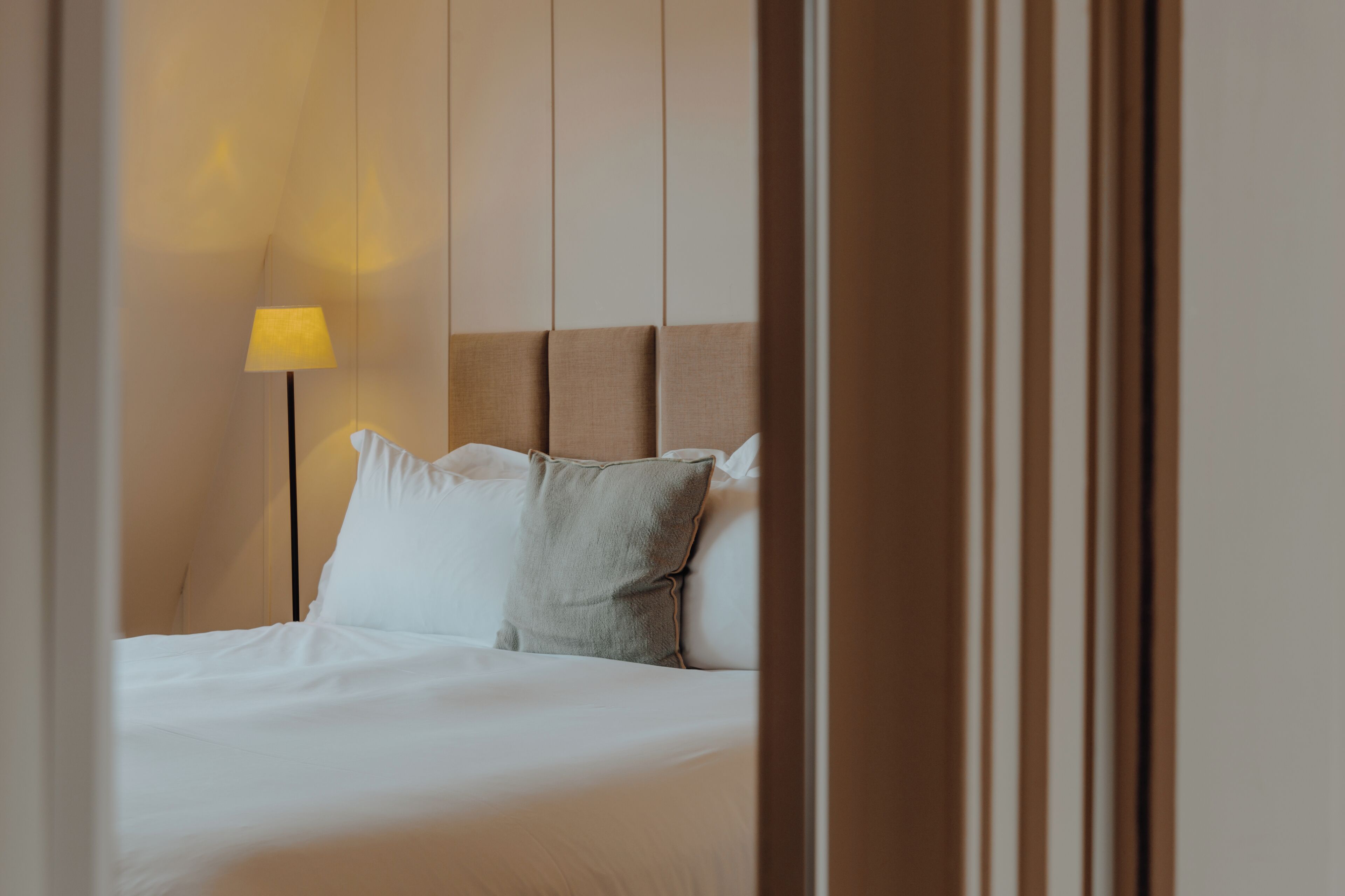 A neatly made bed with white linens and a single grey pillow, a bedside lamp with a yellow shade, and a wooden headboard.