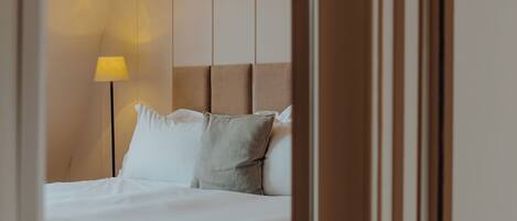 A neatly made bed with white linens and a single grey pillow, a bedside lamp with a yellow shade, and a wooden headboard.