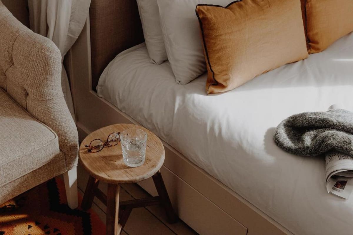 A neatly made bed with white and brown pillows, a wooden stool with a glass of water and glasses, and a rolled-up newspaper on the bed.