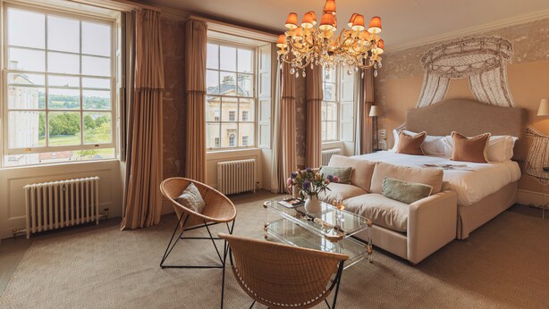 A hotel room with a large bed, a chandelier, a wicker chair, a glass coffee table, and a view of a building through the window.