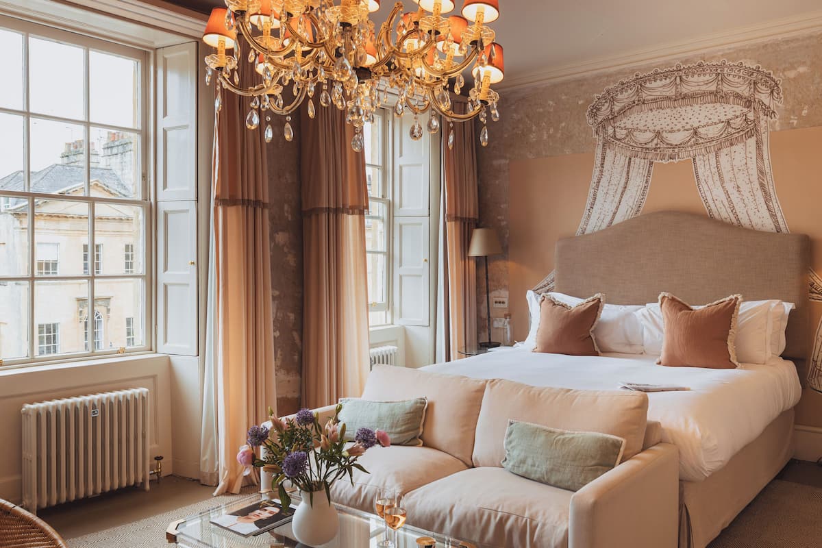 A spacious bedroom with a large bed, a chandelier, and a view of buildings through large windows.
