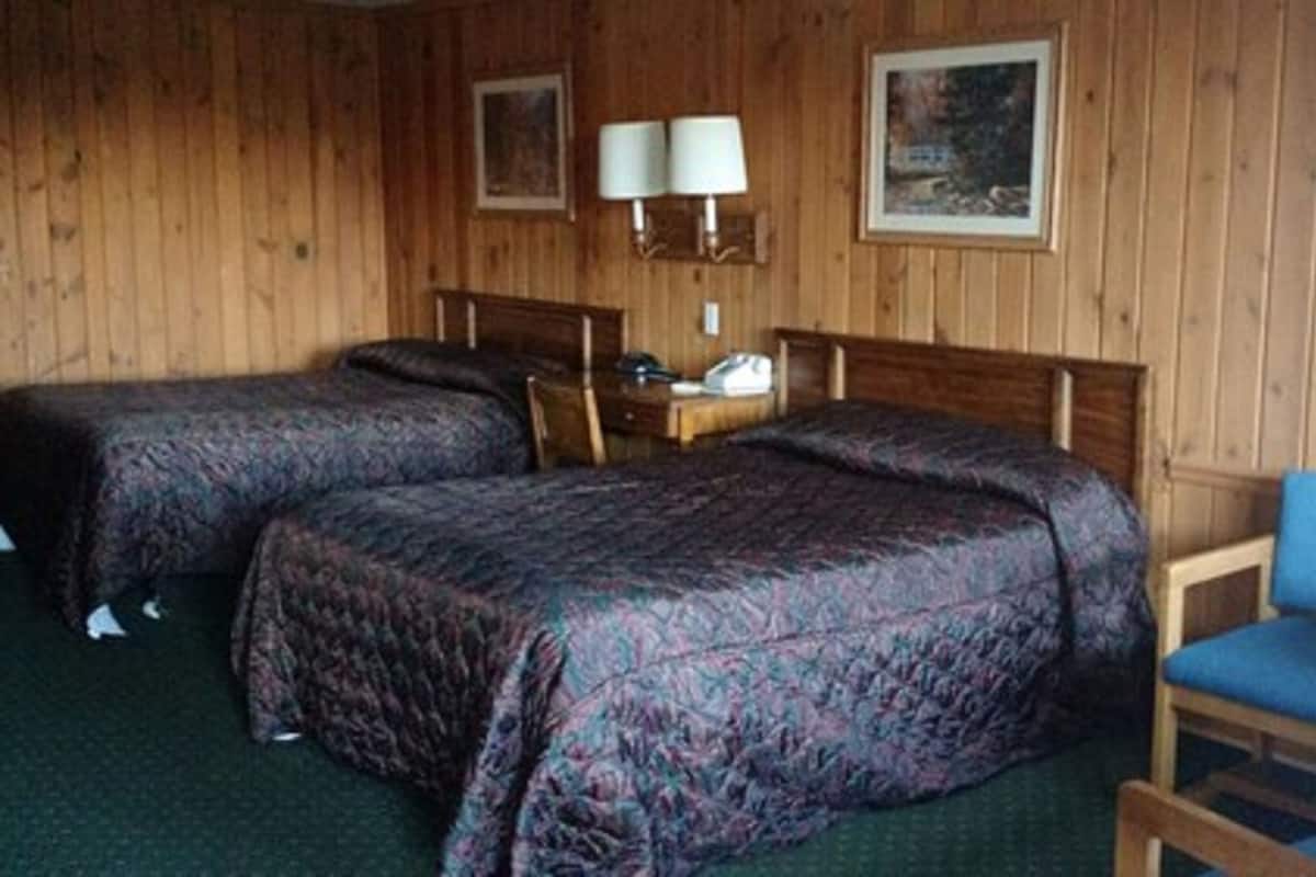 A room with two beds, a chair, a nightstand, and a wall-mounted lamp.