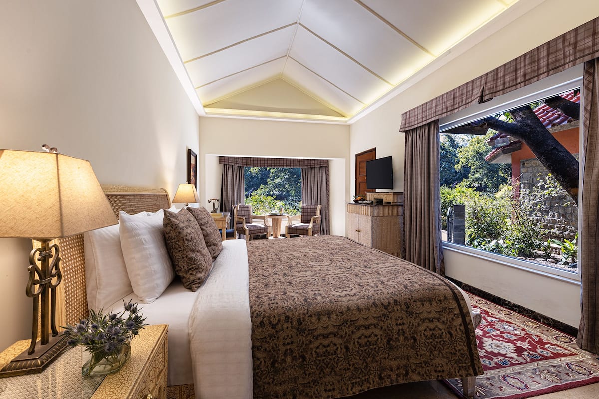 A hotel room with a large bed, two bedside lamps, a TV, a small table with chairs, and a view of the outdoors through a large window.