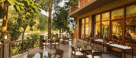 An outdoor dining area with wicker furniture and glass tables, overlooking a river and lush greenery.