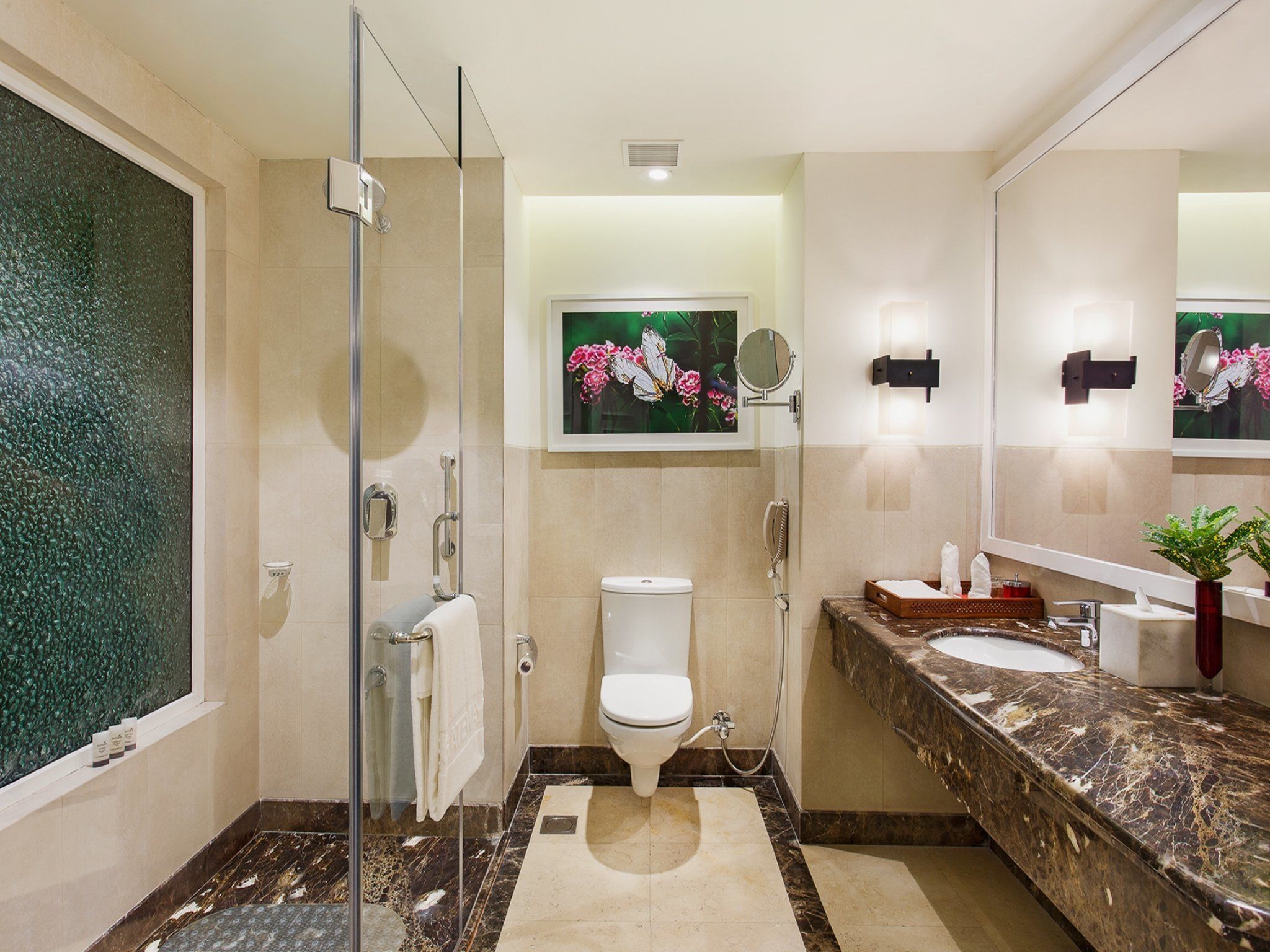 A modern bathroom with a glass shower enclosure, a toilet, a sink with a marble countertop, and a mirror with a flower painting above it.