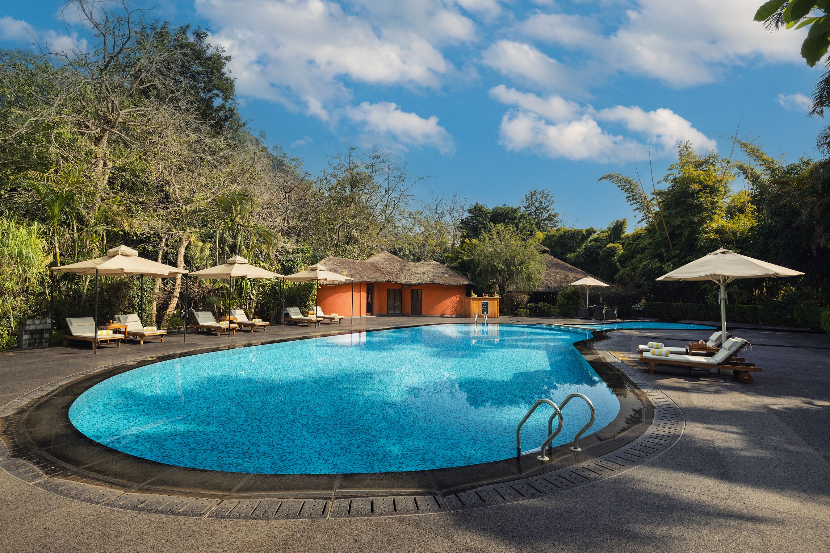 A swimming pool with lounge chairs and umbrellas, surrounded by trees and a thatched-roof building.