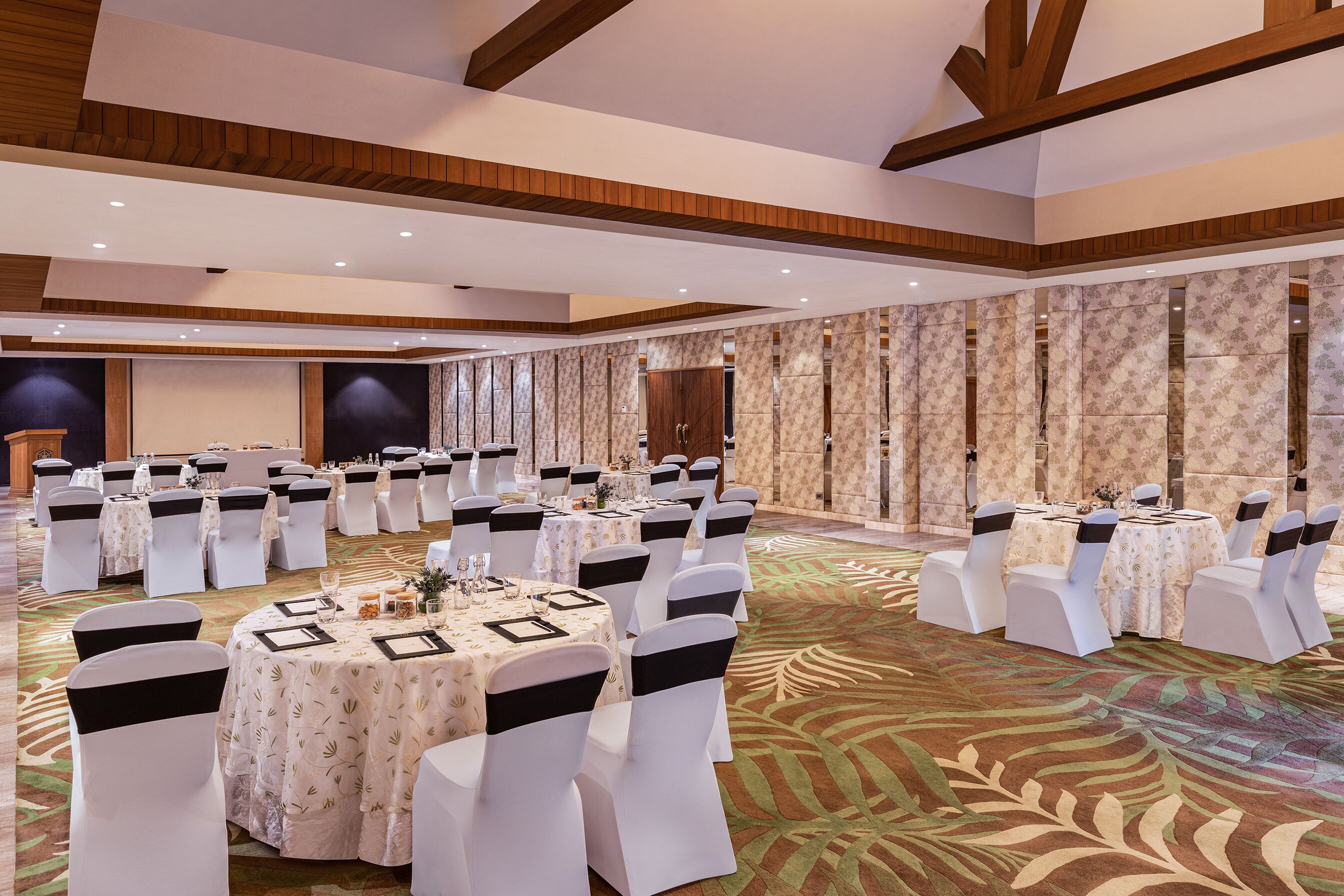 Un salón de banquetes con mesas redondas, sillas blancas adornadas con cintas negras y una alfombra estampada.