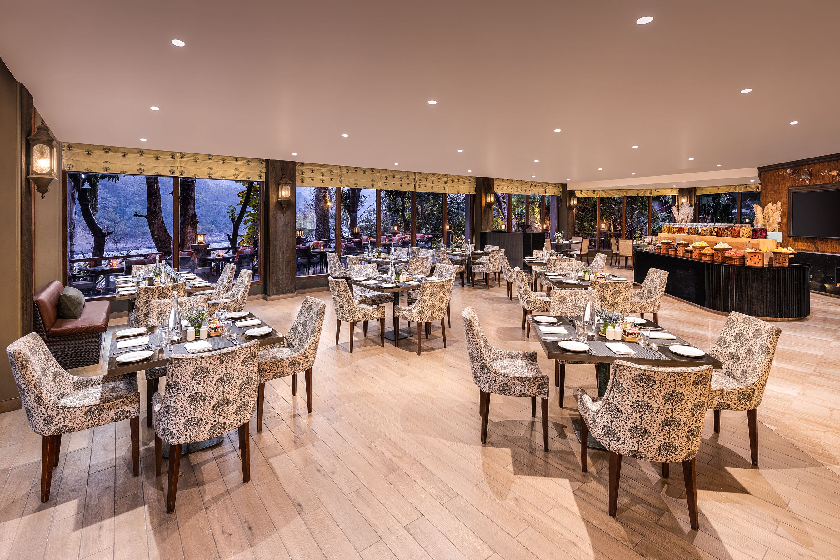 Un comedor amplio con varias mesas y sillas, ventanas grandes que ofrecen vistas a árboles y una masa de agua, y un mostrador de buffet con diversos alimentos.