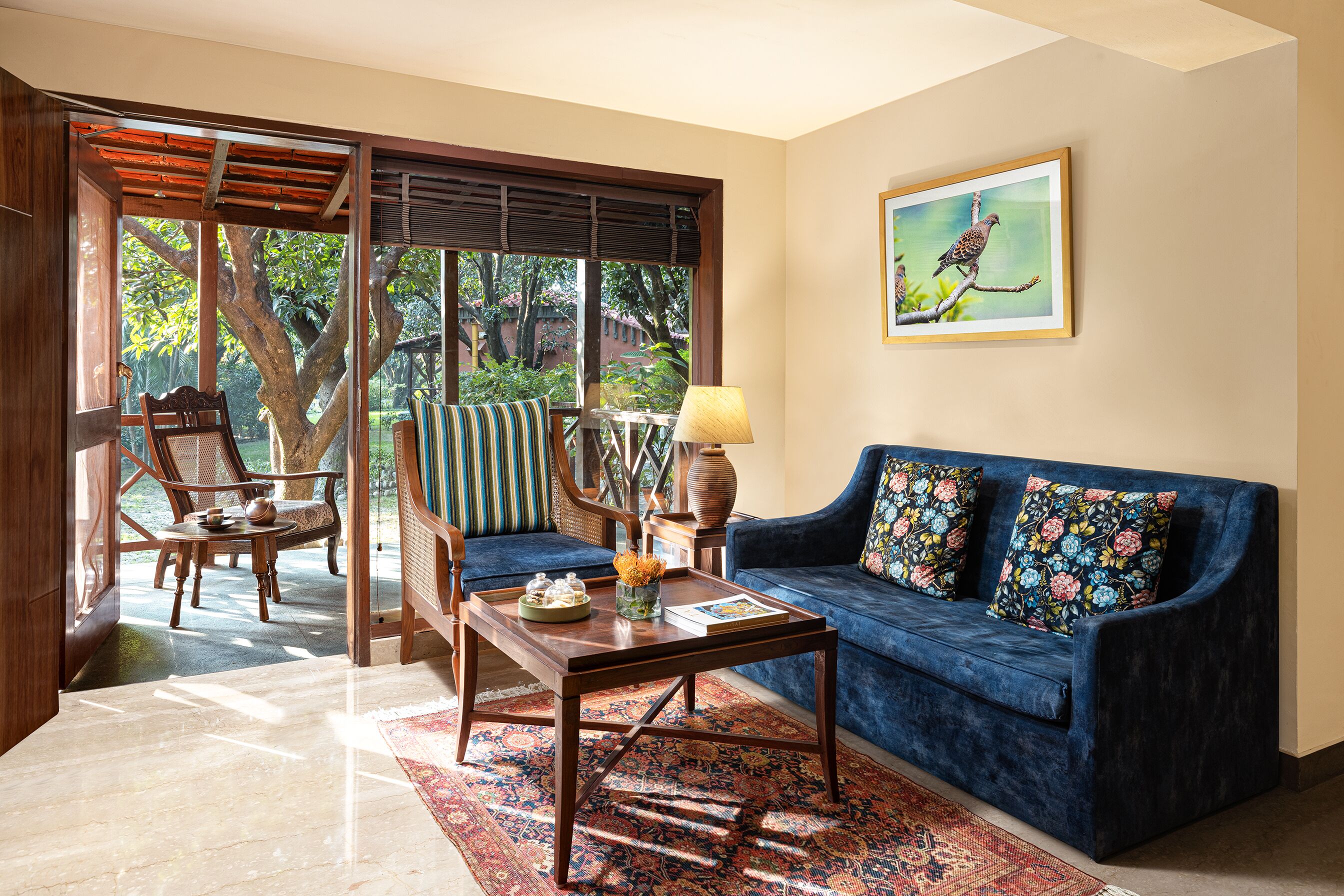 A living room with a blue sofa, a wooden coffee table, and a chair. There is a window with a view of trees and a painting on the wall.