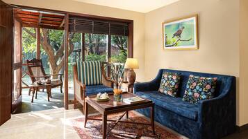 A living room with a blue sofa, a wooden coffee table, and a chair. There is a window with a view of trees and a painting on the wall.