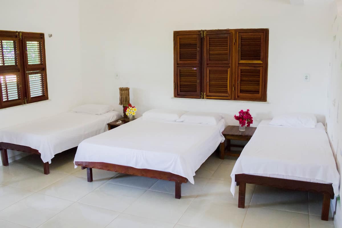 Una habitación con tres camas individuales, una mesita de noche de madera con un florero y una ventana con contraventanas de madera.