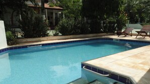 Una piscina con borde de azulejos, rodeada de un jardín con diversas plantas y un edificio al fondo.