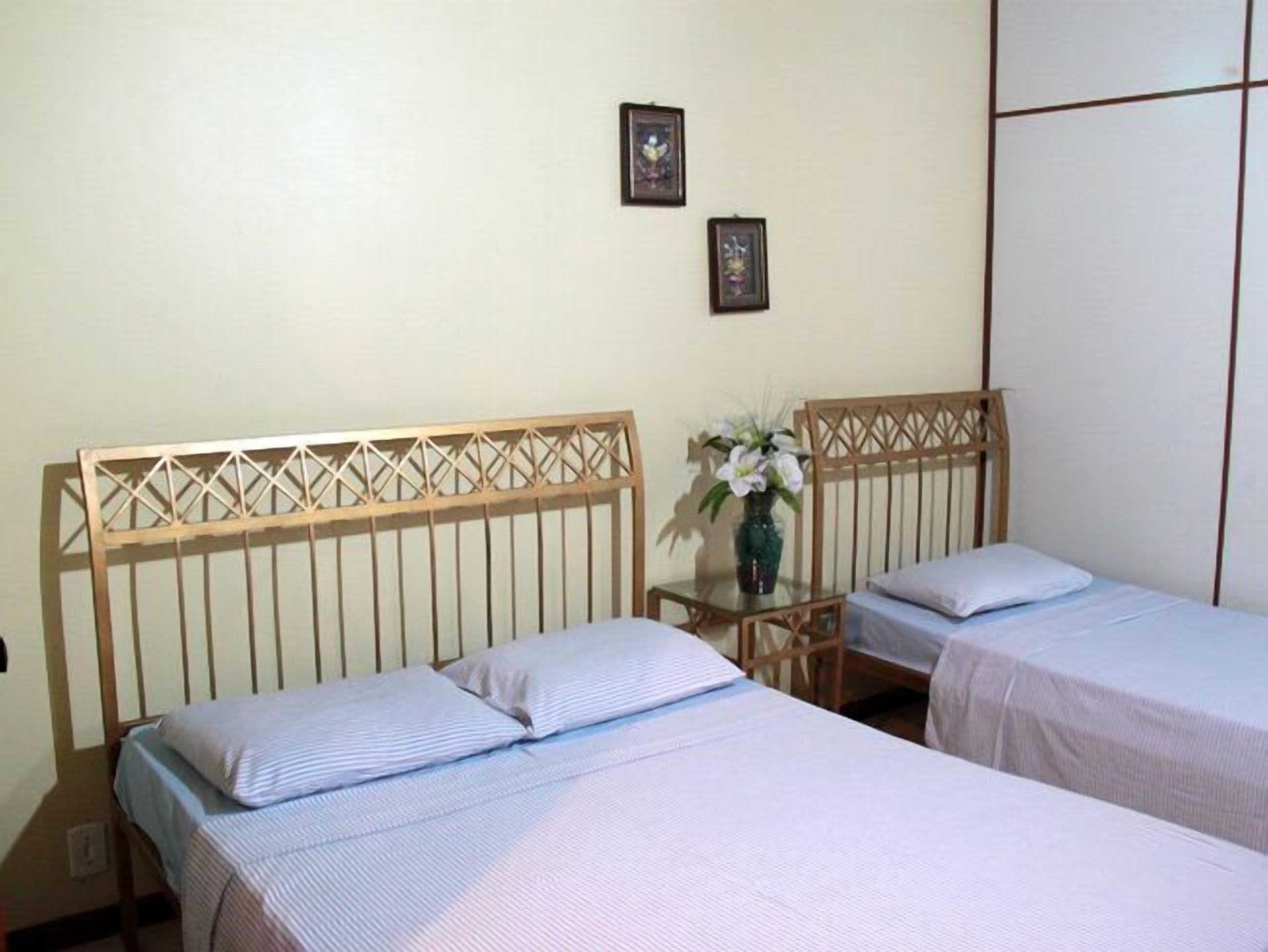 A room with two single beds with wooden headboards, a small table with a vase of flowers, and two framed pictures on the wall.