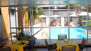 An indoor pool area with tables and chairs, a stone wall, and a view of the outdoor pool. - Pousada Praia da Costa (Vila Velha)
