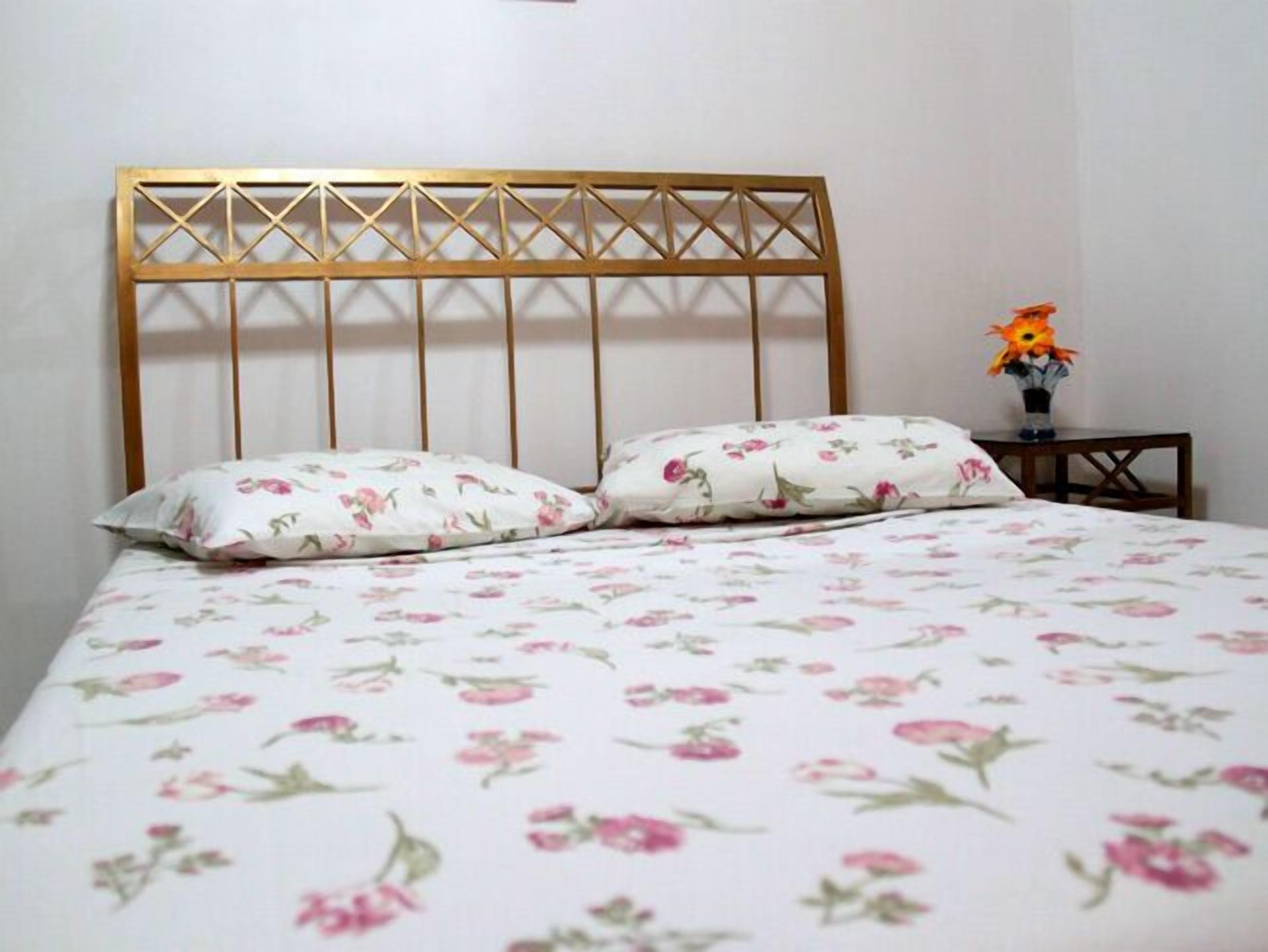 A bed with a floral-patterned bedspread and two matching pillows, a wooden headboard, and a small table with a vase of flowers.