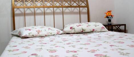 A bed with a floral-patterned bedspread and two matching pillows, a wooden headboard, and a small table with a vase of flowers.