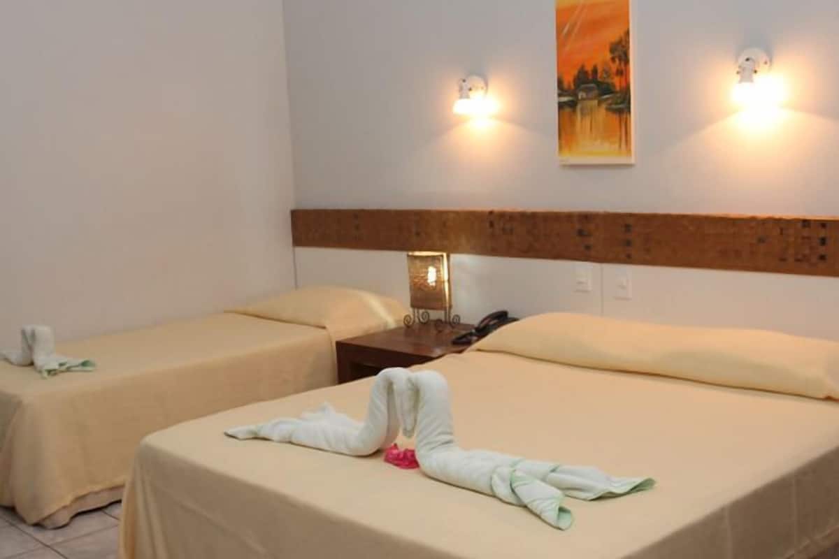 Habitación de hotel con dos camas, cada una con colcha y almohadas color beige. Entre las camas, una mesita de noche con lámpara y teléfono. Un cuadro cuelga de la pared sobre las camas.
