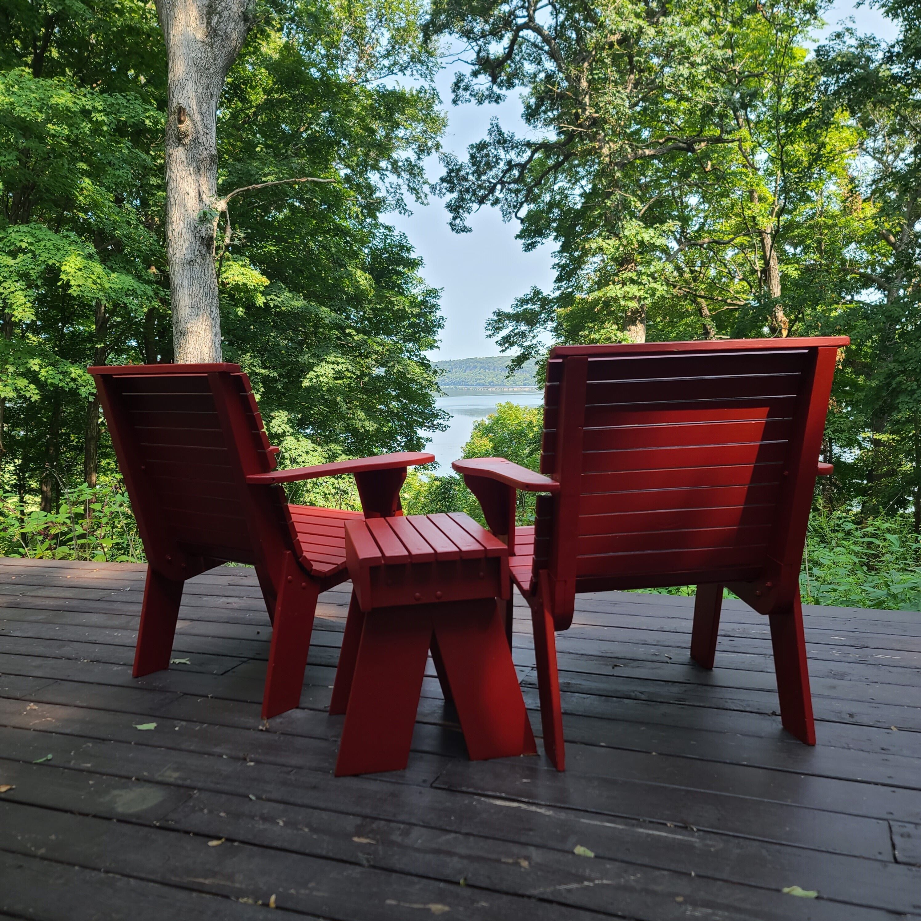 Deux chaises Adirondack rouges sont posées sur une terrasse en bois, d’où l’on peut admirer un lac et des arbres.