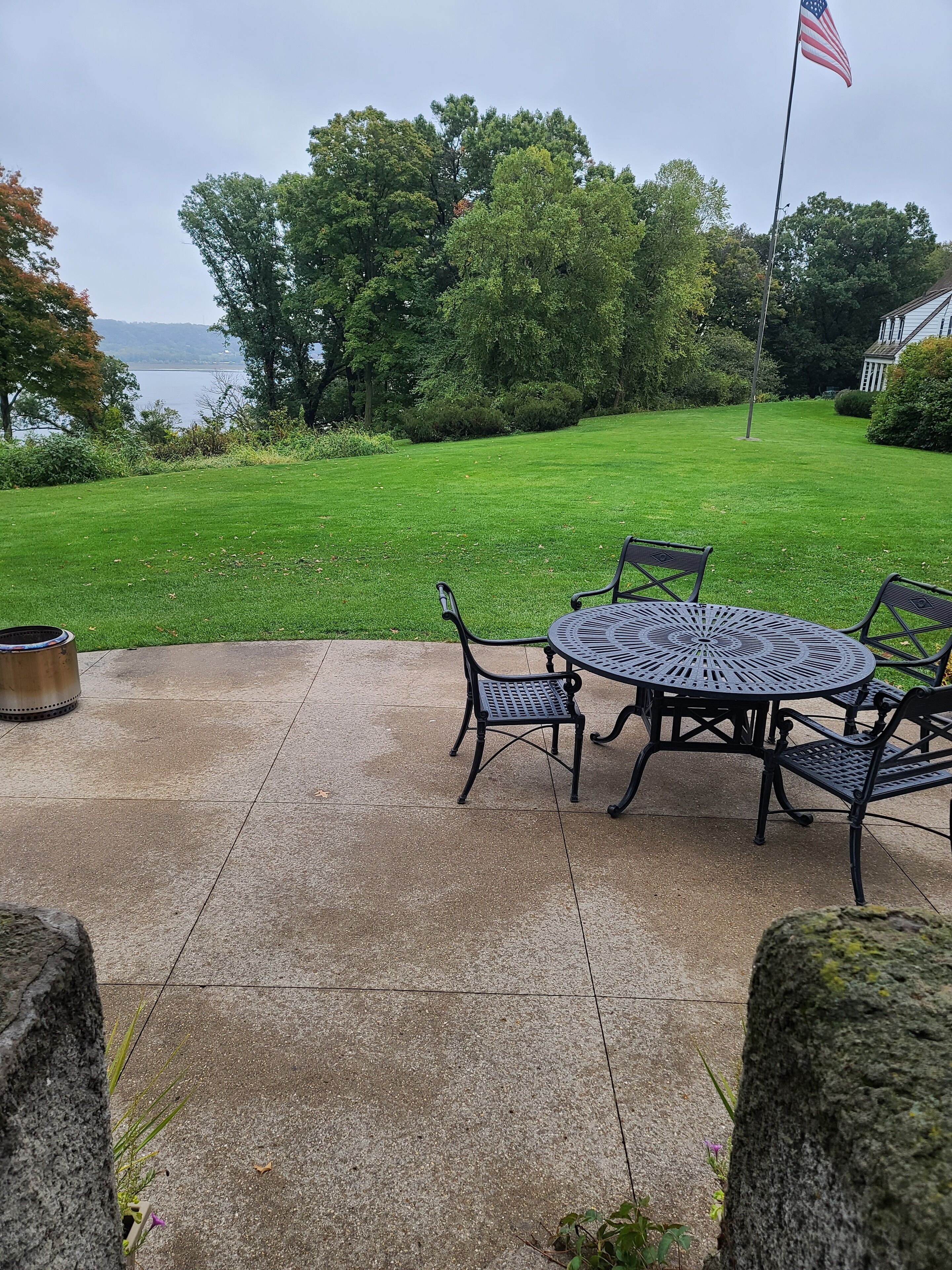 Eine Terrasse mit rundem Tisch und Stühlen, einer Flagge und Blick auf einen See und Bäume.