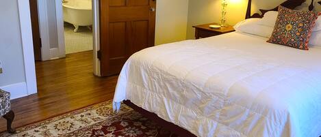 A bedroom with a large bed, a wooden door, a nightstand with a lamp, and a patterned rug.