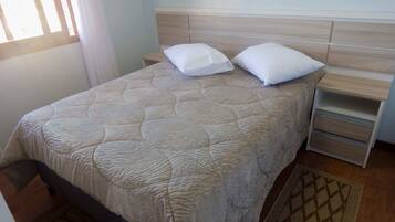 A neatly made bed with a quilted headboard, a bedside table with drawers, and a patterned rug on the floor.