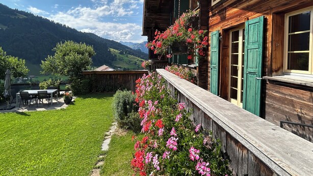 A wooden chalet with a balcony overlooking a lush garden and a mountainous landscape.