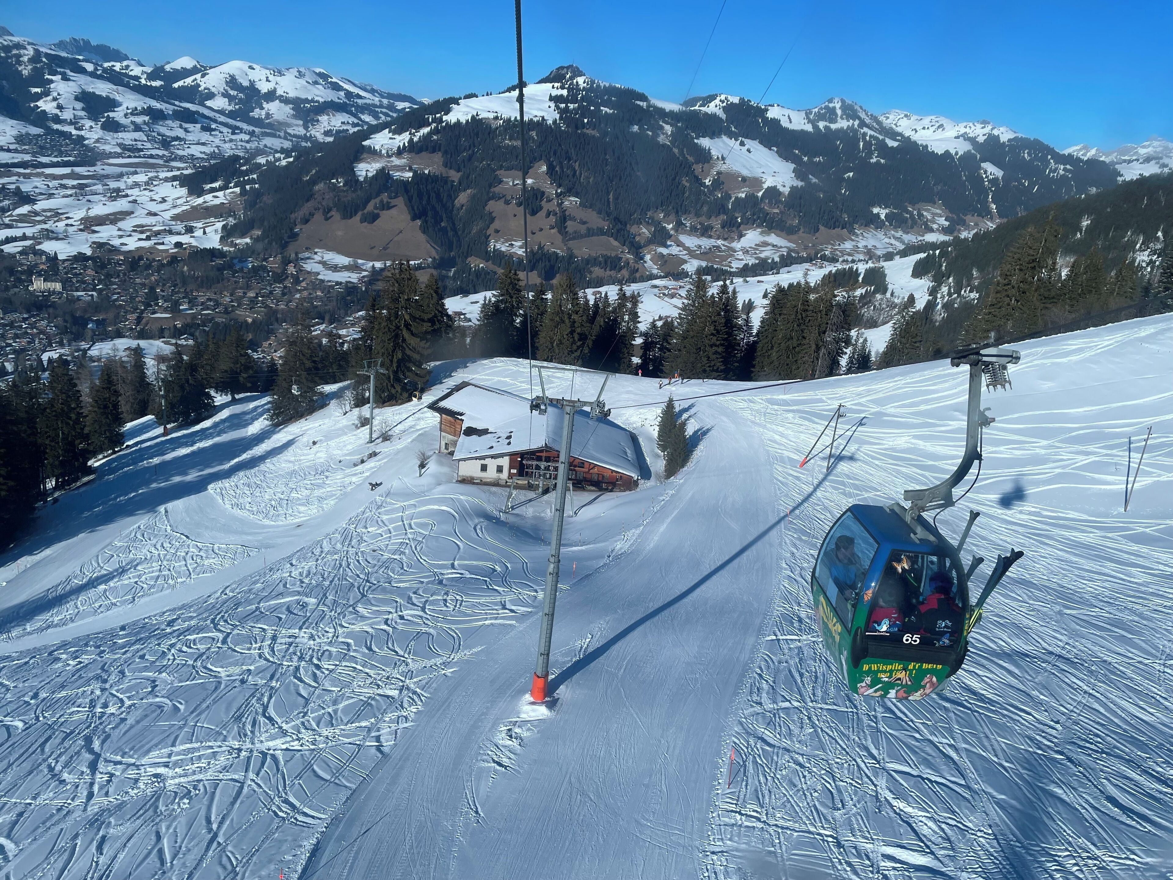 Un asiento de telesilla deslizándose por una ladera nevada, con montañas al fondo y un pequeño edificio con tejado rojo.
