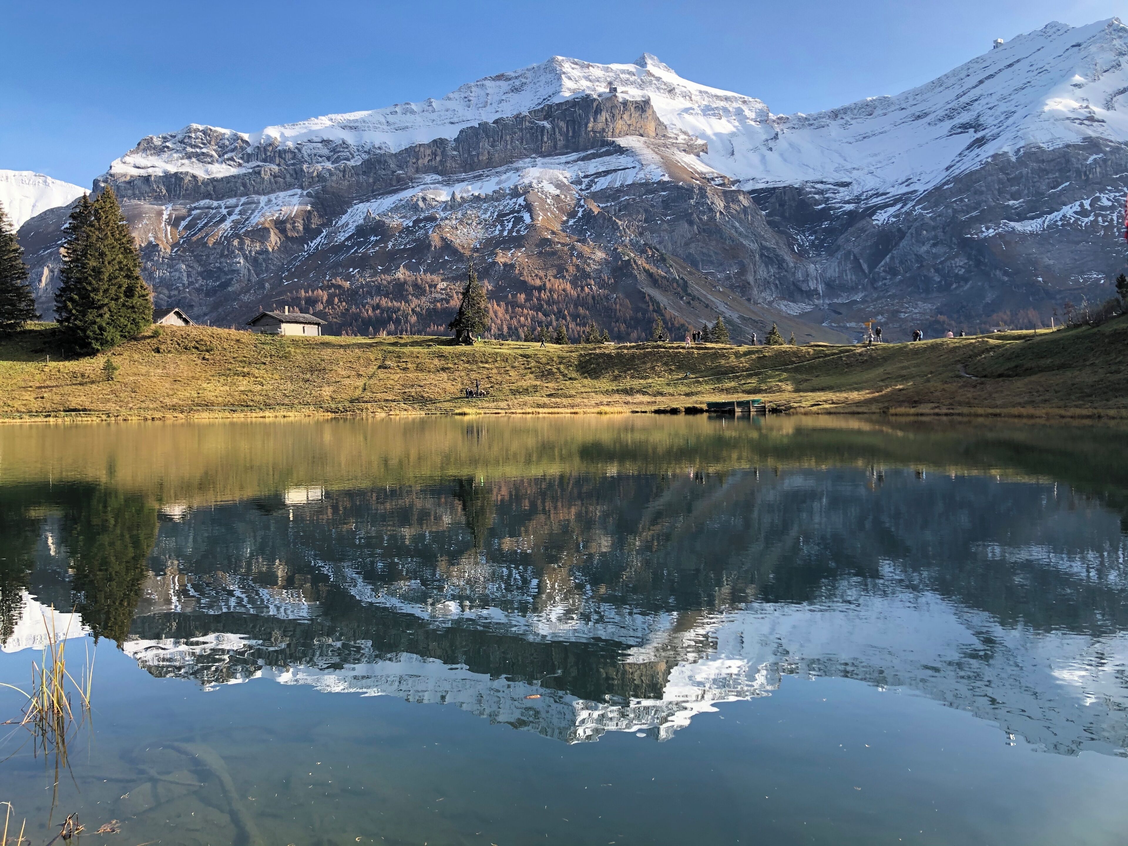 A serene mountain landscape with a clear lake reflecting the surrounding peaks and a few buildings on the hillside.