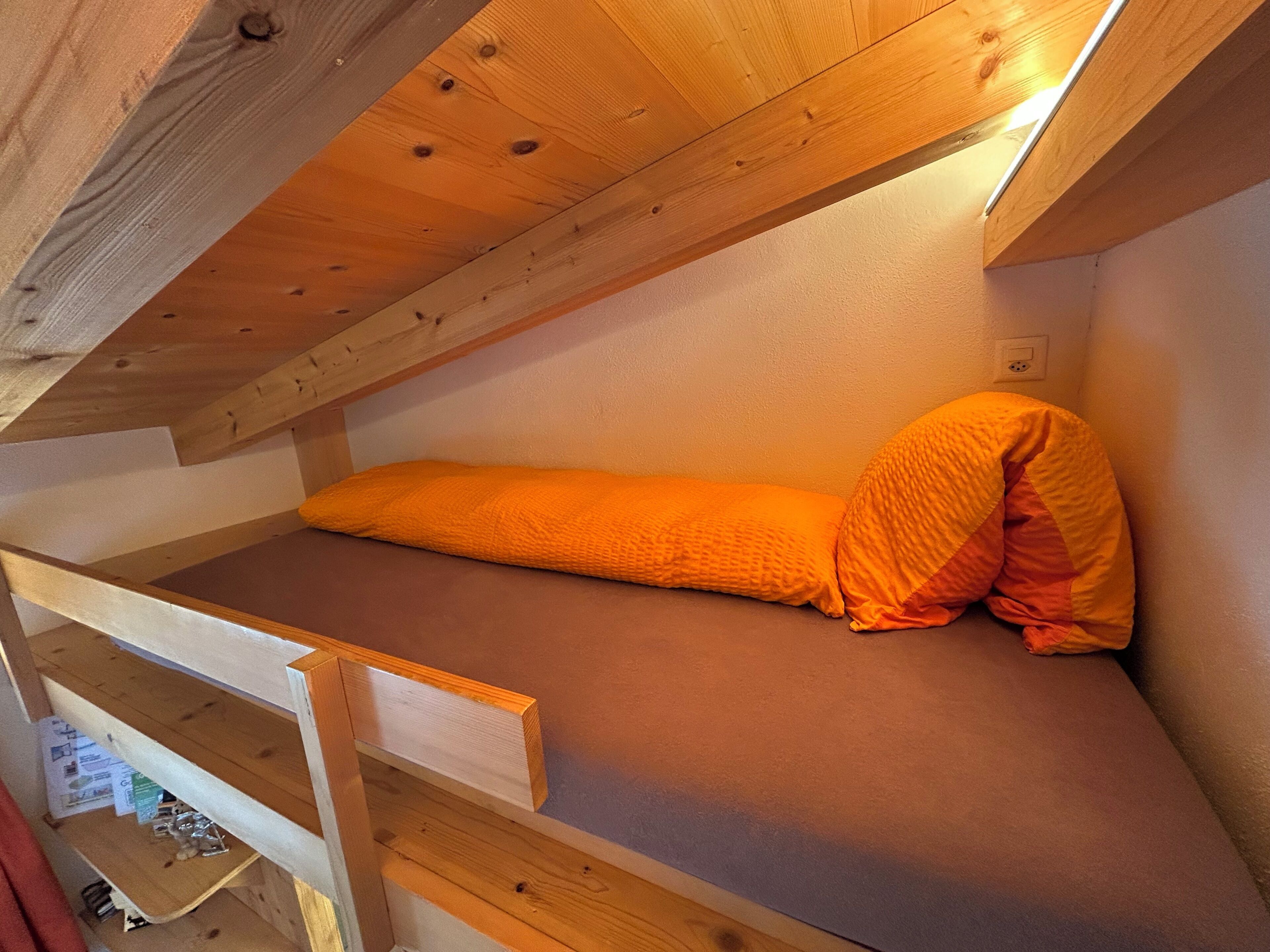 A bunk bed with an orange mattress and pillows, wooden beams, and a sloped ceiling.