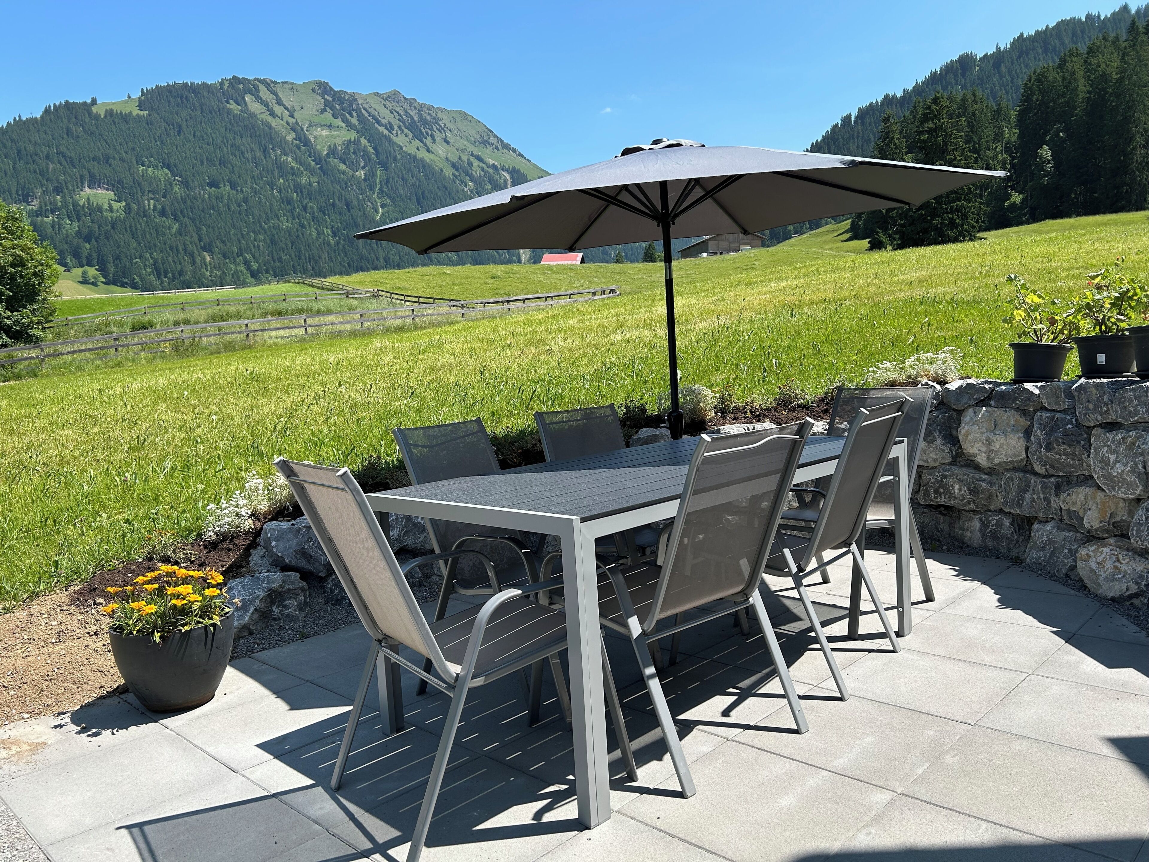 En udendørs terrasse med et bord, stole, parasol og udsigt over et grønt område og bjerge.