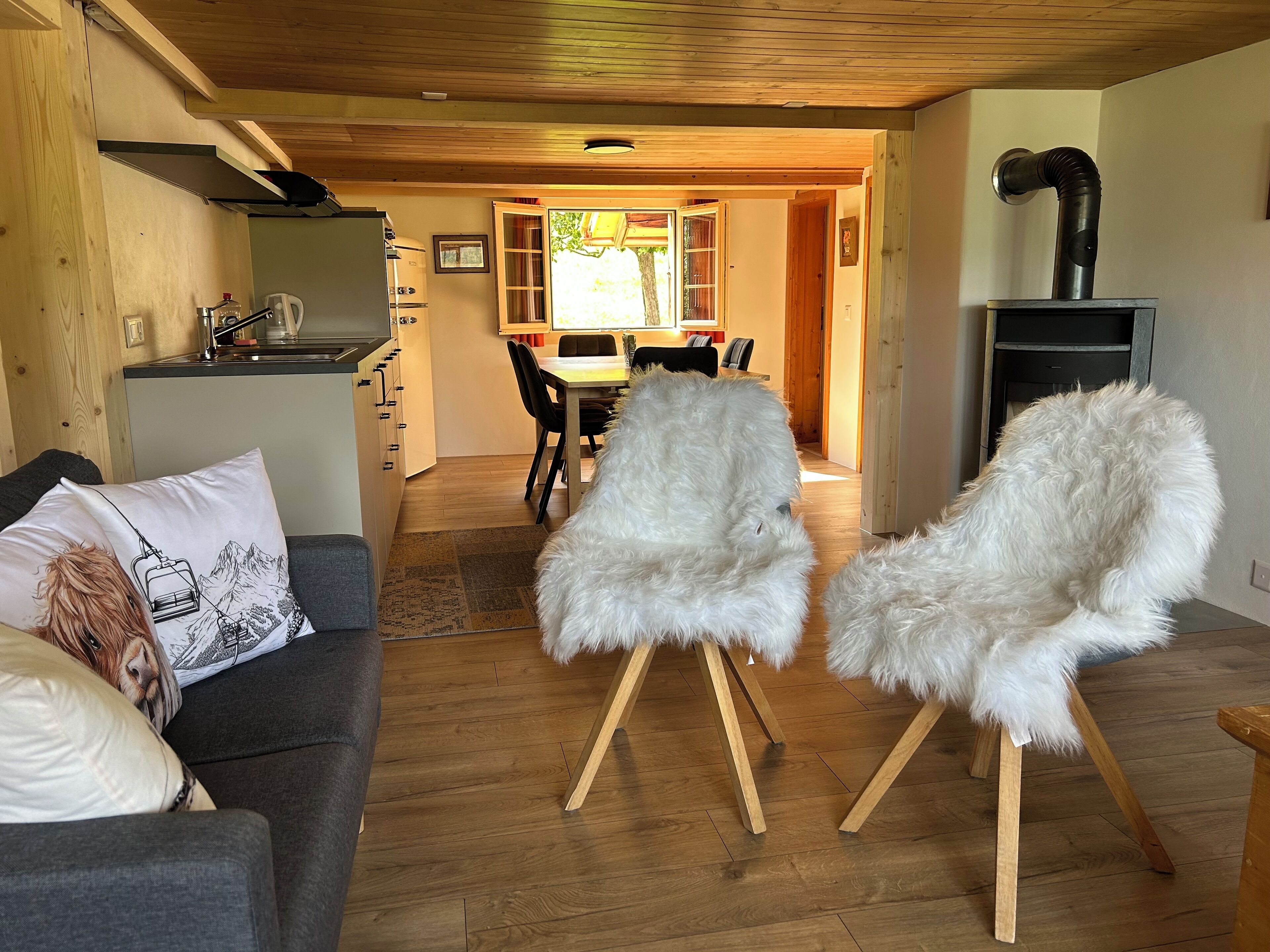 A cozy living room with a grey sofa, two white furry chairs, a wooden ceiling, and a kitchen area in the background.