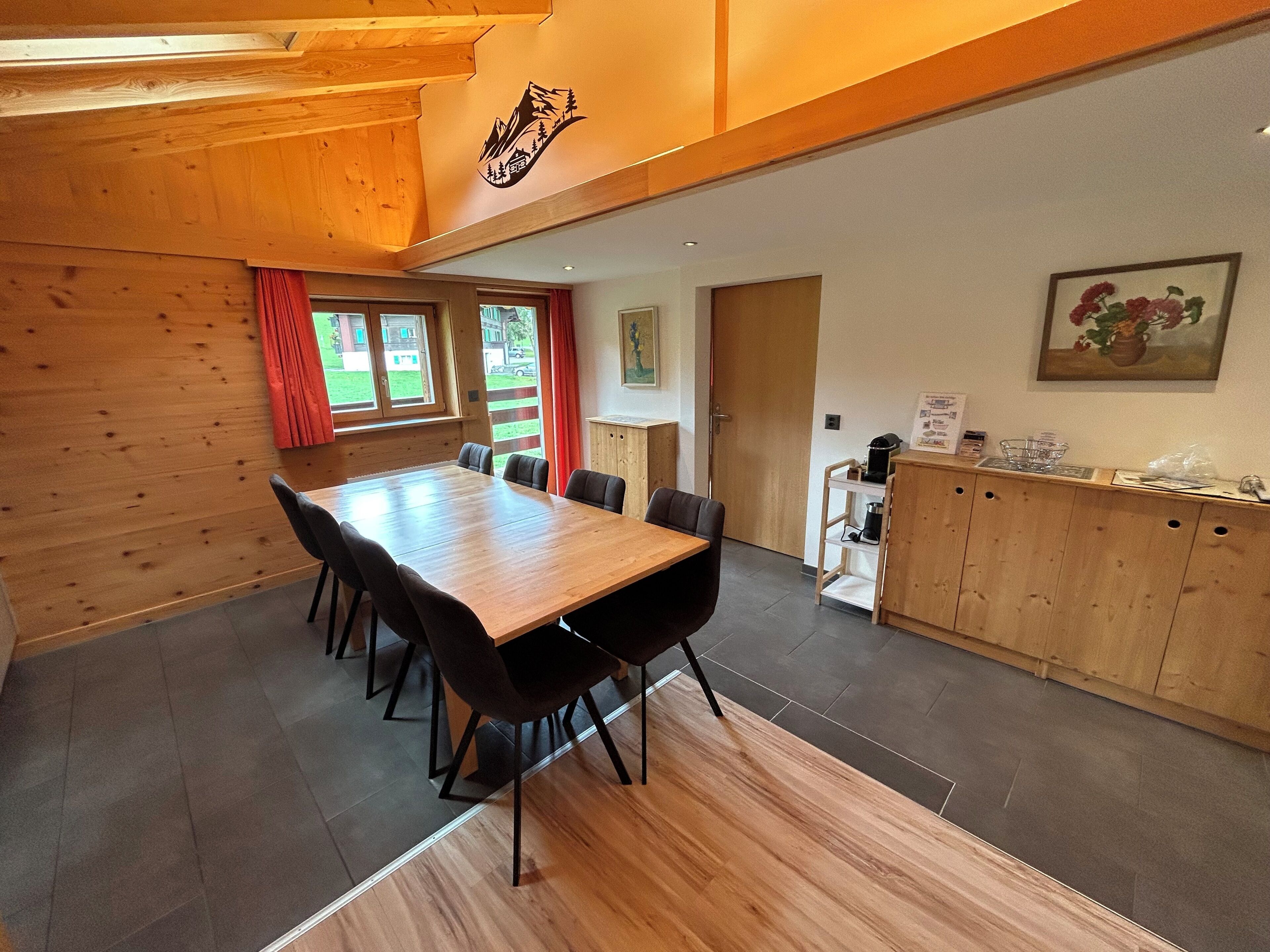 Un comedor de madera con una mesa larga y sillas, una zona de cocina con armarios, y un cuadro en la pared.