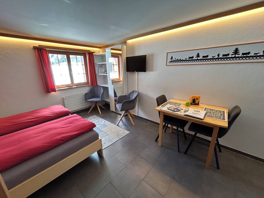 A room with a bed, a desk, two chairs, a TV, and a window with curtains.