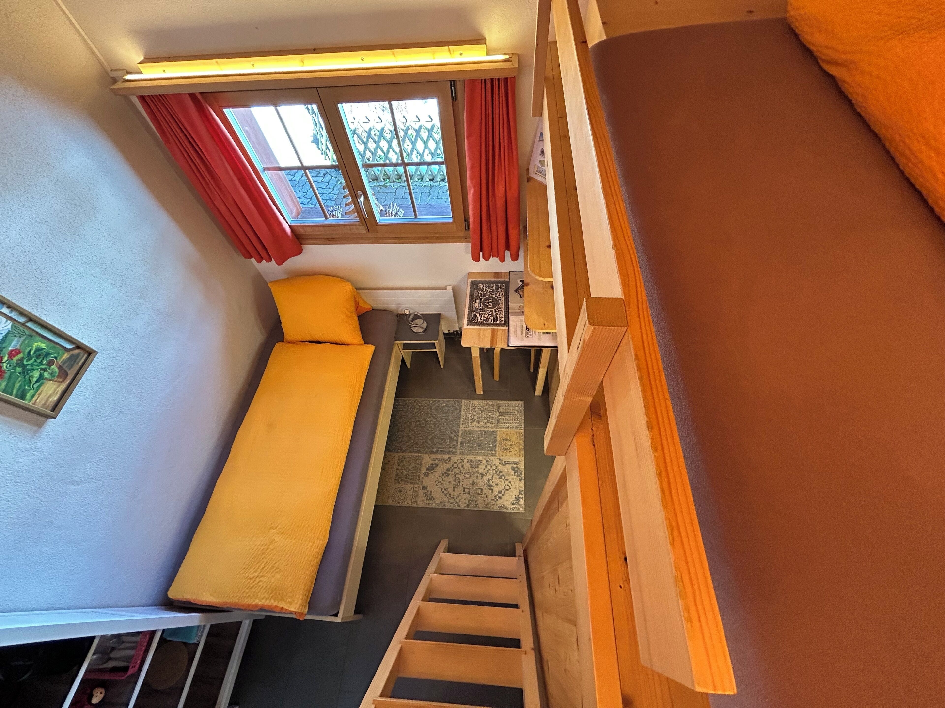 Una habitación tipo dormitorio con cama, una mesita, una silla, un ventanal con cortinas y una escalera.