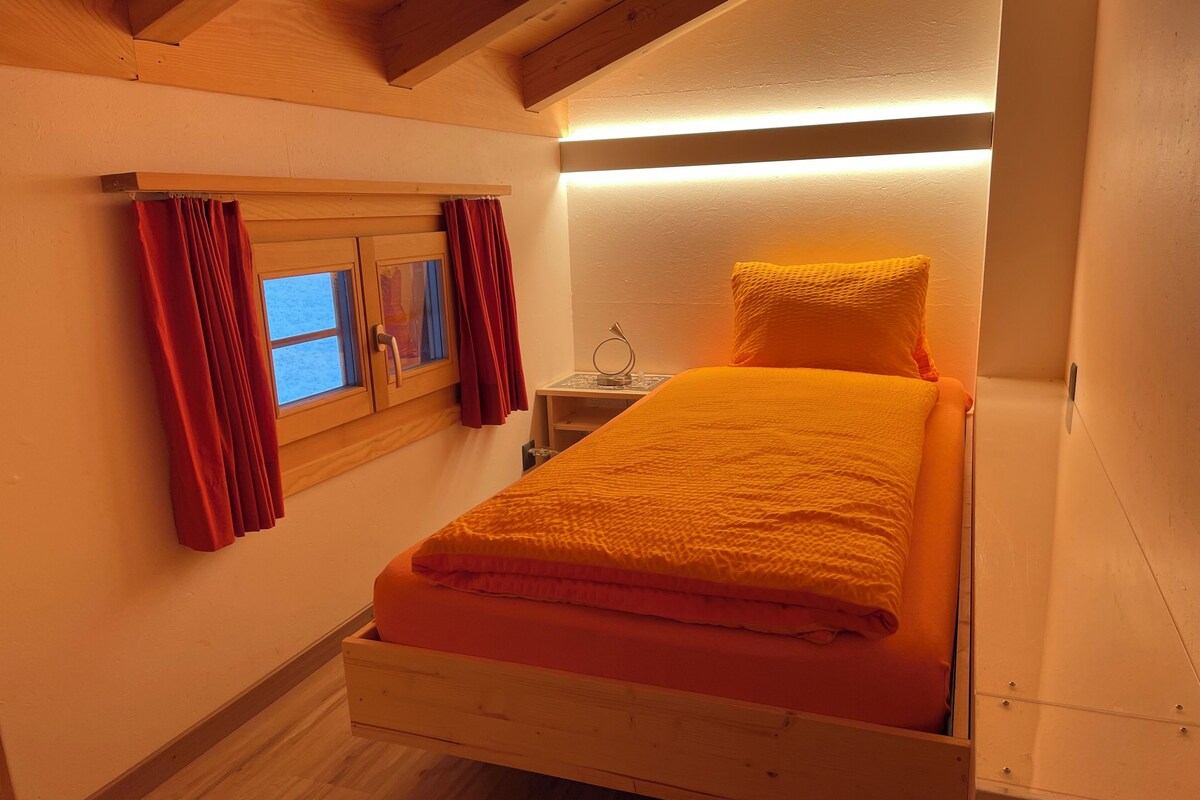 A small, cozy bedroom with a wooden bed, a window with curtains, and a bedside table.