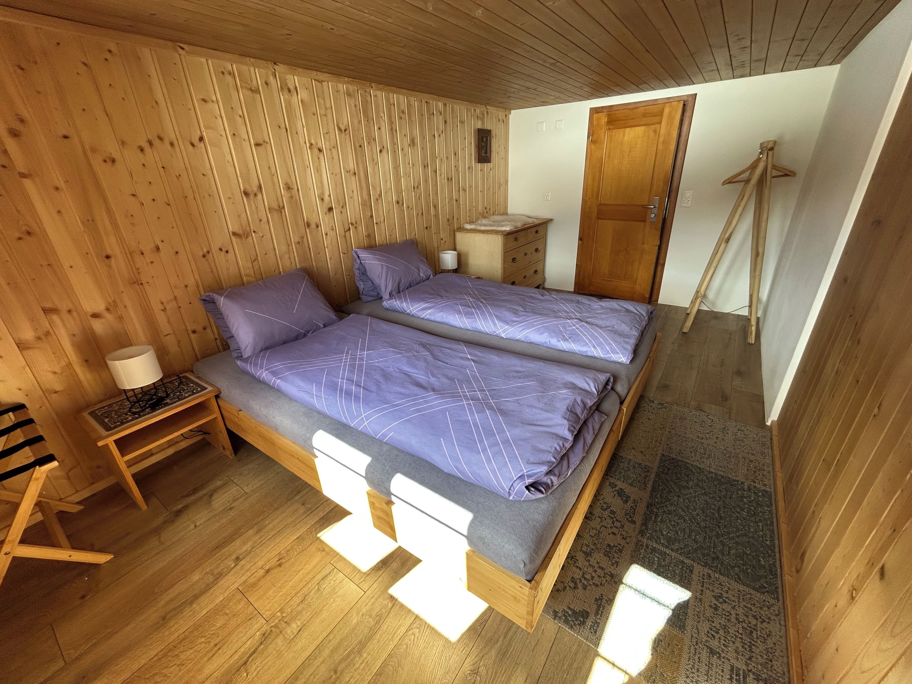 A wooden-paneled room with two beds, a nightstand, a wooden chair, and a door.
