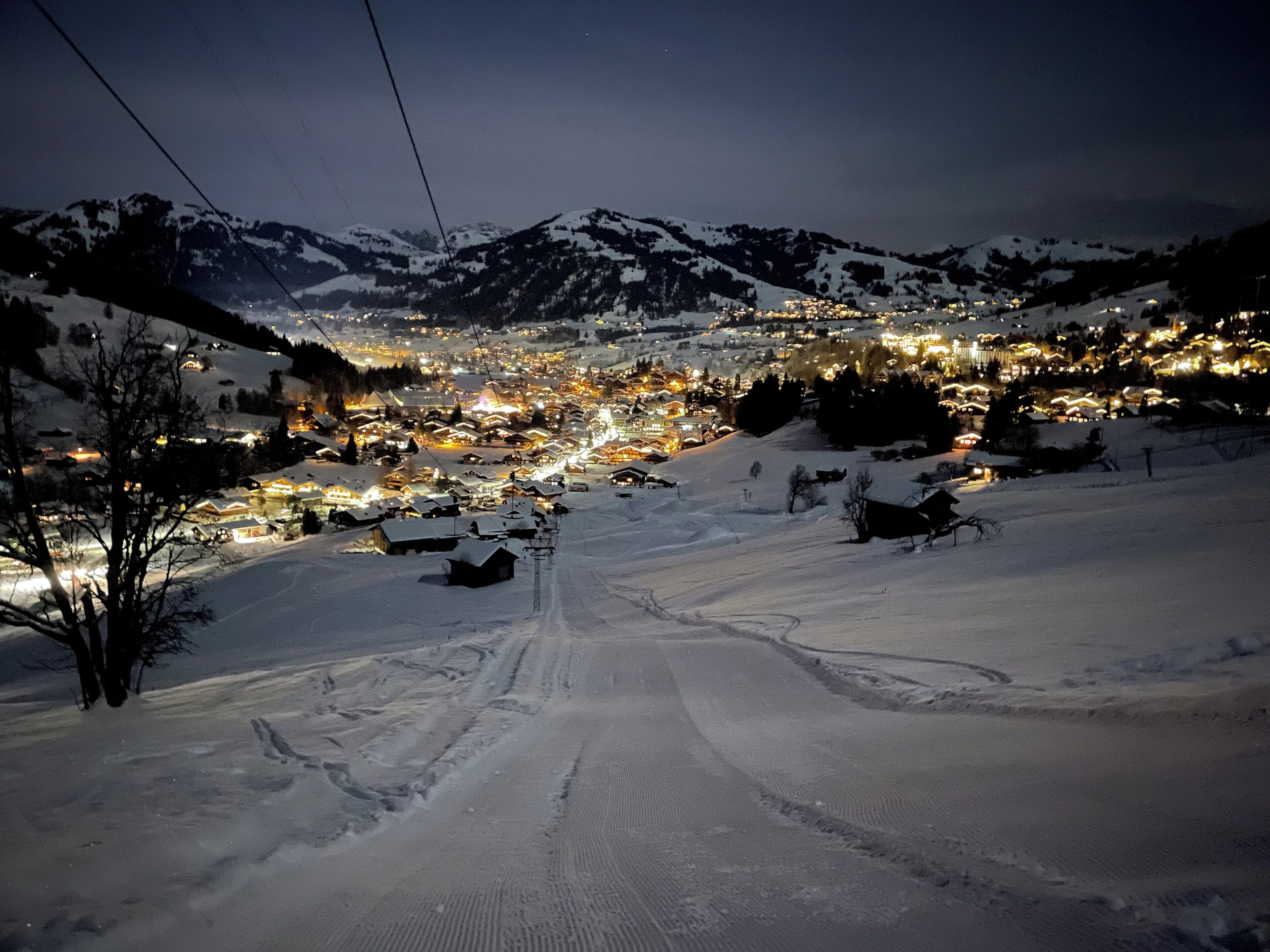 A snow-covered village with illuminated buildings, a clear sky, and a ski slope.