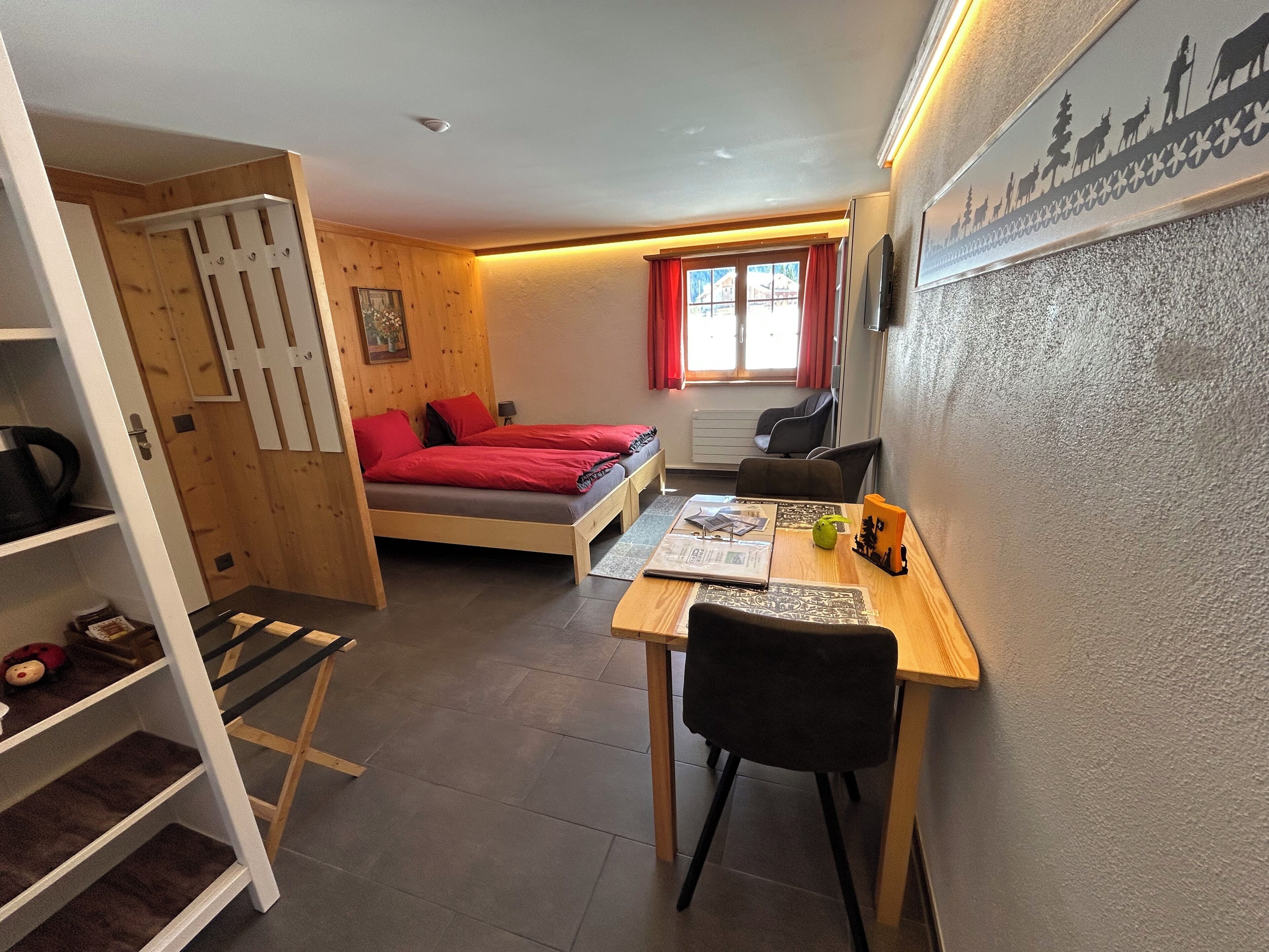 A compact room with a bed, a desk, and a chair. There is a window with red curtains, a radiator, and a shelf with items.