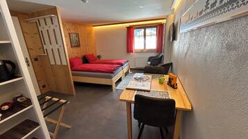 A compact room with a bed, a desk, and a chair. There is a window with red curtains, a radiator, and a shelf with items.